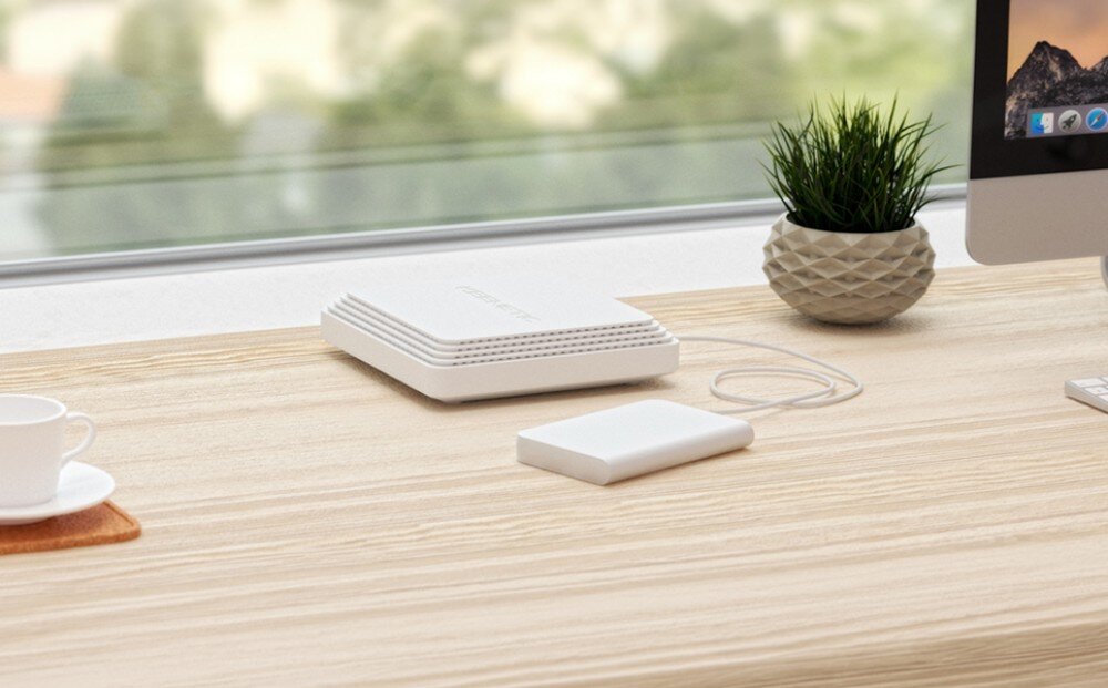 Router KEENETIC Hopper SE Białe, płaskie urządzenie bez zewnętrznych anten leży na drewnianym blacie obok białego dysku zewnętrznego, a w tle widać monitor komputera i okno