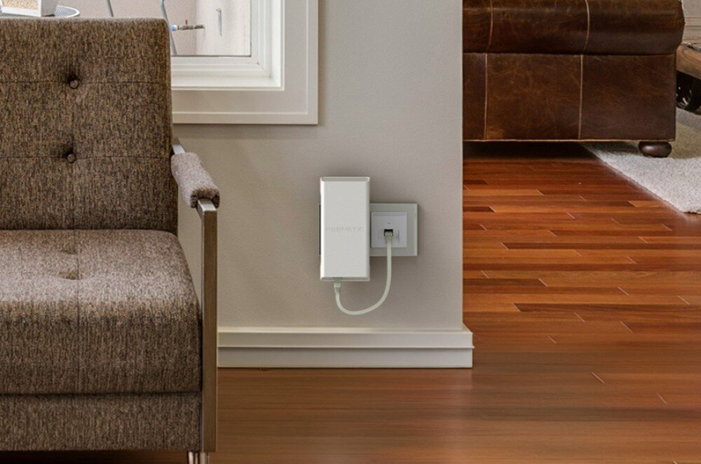 Wzmacniacz sieci KEENETIC Buddy 6 SE Białe prostokątne urządzenie zamontowane na ścianie i połączone kablem z gniazdkiem, obok brązowy fotel i drewniana podłoga