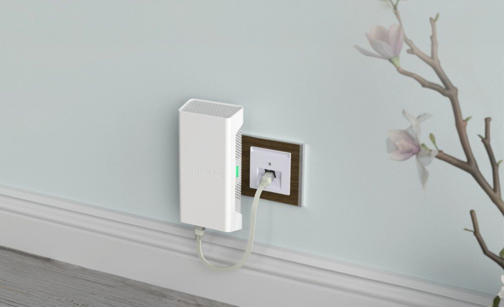 Wzmacniacz sieci KEENETIC Buddy 6 SE Białe urządzenie wpięte kablem do gniazdka w brązowej ramce na błękitnej ścianie, obok gałązka z jasnymi kwiatami