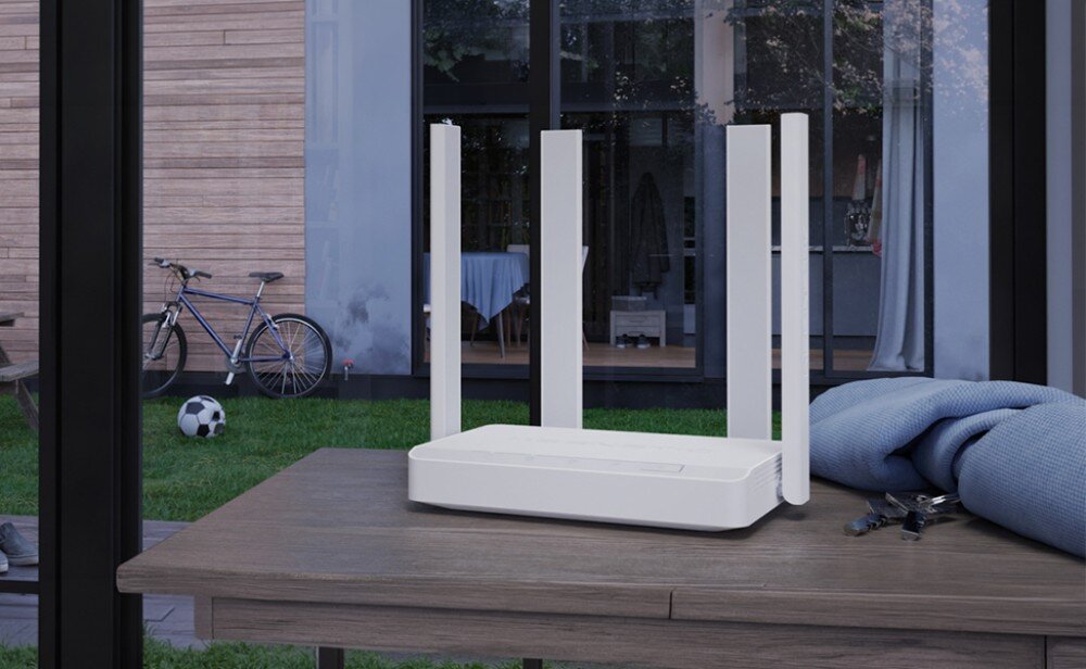 Router KEENETIC Explorer 4G Biały router z czterema antenami na drewnianym stole na tle przeszklonych drzwi, przez które widać ogród z rowerem i piłką