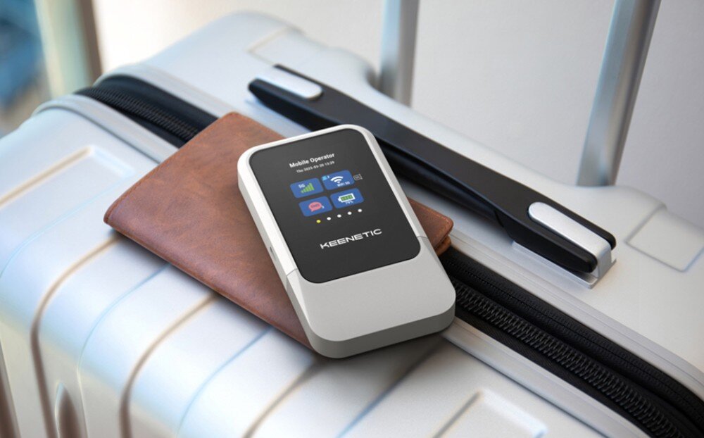 Router KEENETIC Roamer 5G Przenośny router o srebrnej obudowie z czarnym ekranem leży na brązowym portfelu, który umieszczono na zamkniętej, metalicznej walizce podróżnej