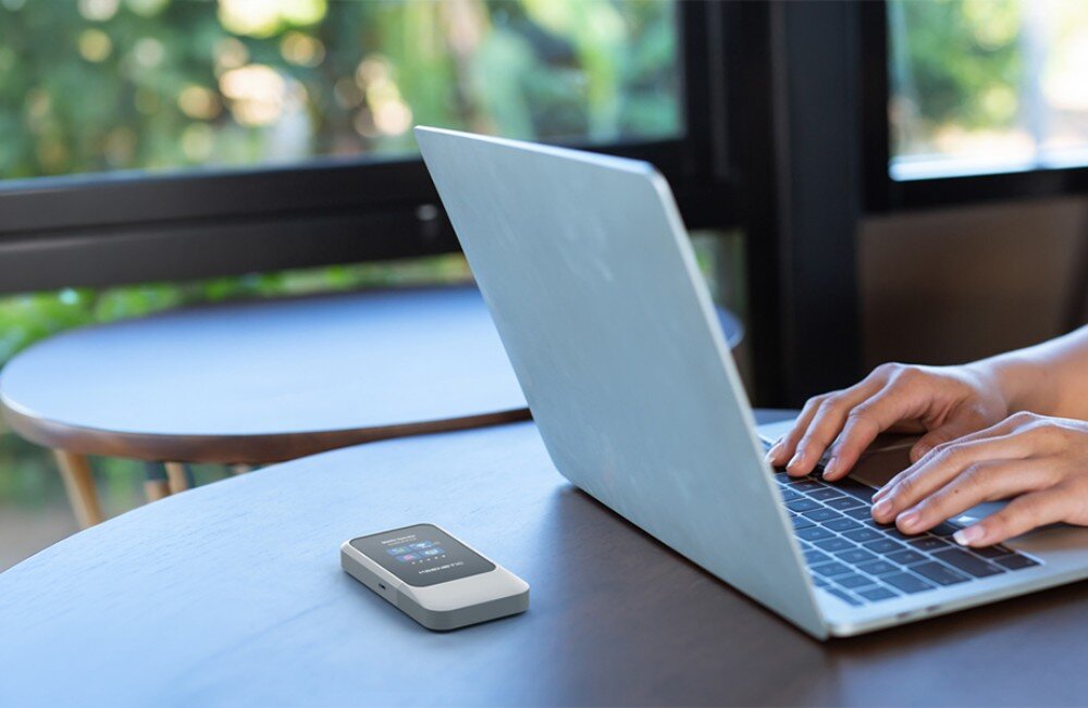 Router KEENETIC Roamer 5G Mały router bezprzewodowy z kolorowymi ikonami na wyświetlaczu spoczywa na drewnianym stoliku obok otwartego laptopa, na którym pracuje osoba w kawiarni