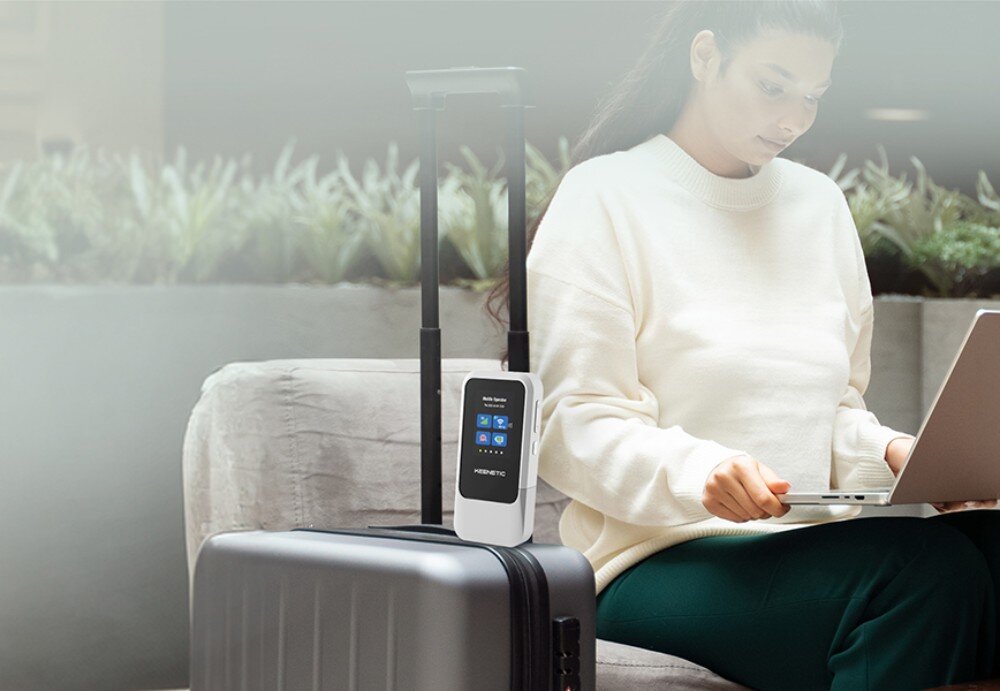 Router KEENETIC Roamer 5G Kobieta siedząca z laptopem na kolanach przy dużym bagażu podróżnym, na którym ustawiono pionowo jasne urządzenie sieciowe z logotypem Keenetic