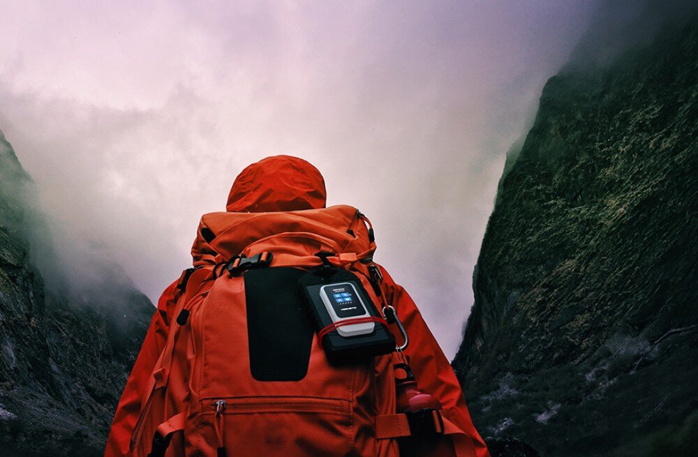 Router KEENETIC Roamer 5G Turysta w czerwonej kurtce z dużym plecakiem idzie przez zamglony, górzysty teren, mając przypięty do tylnej części bagażu mobilny router w czarnym etui
