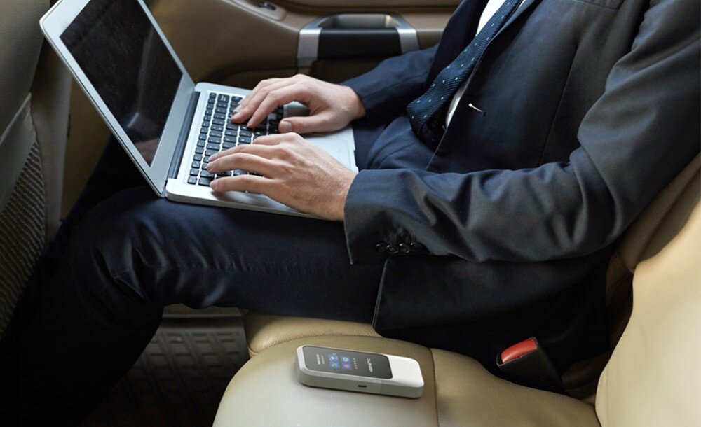 Router KEENETIC Roamer 5G Mężczyzna w garniturze pracuje na laptopie podczas jazdy na tylnym siedzeniu samochodu, a obok niego na jasnej tapicerce leży płaskie urządzenie elektroniczne