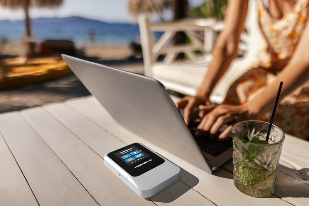 Router KEENETIC Roamer 5G Urządzenie mobilne z ekranem wskazującym status połączenia spoczywa na białym, drewnianym stole na plaży obok szklanki z napojem i osoby piszącej na komputerze