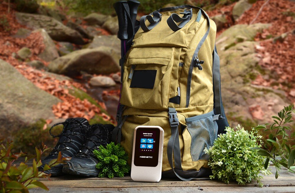 Router KEENETIC Roamer 5G Kieszonkowy router stoi pionowo na drewnianym podłożu w otoczeniu ekwipunku turystycznego, butów trekkingowych oraz beżowego plecaka w leśnej scenerii