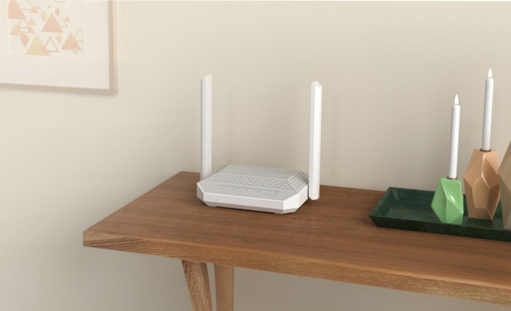 Router KEENETIC Racer Biały router z dwiema pionowymi antenami stoi na brązowym, drewnianym stole w jasnym pokoju, obok kolorowych świeczników i obrazka na ścianie