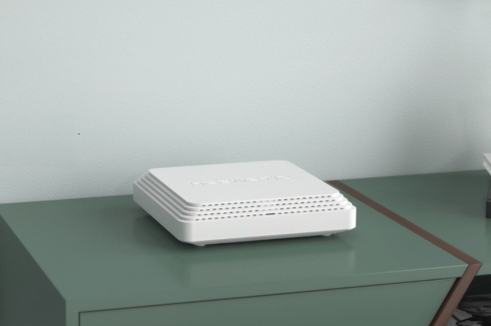 Router KEENETIC Challenger SE Białe kwadratowe urządzenie z poziomymi nacięciami na bokach leżące na zielonym blacie mebla