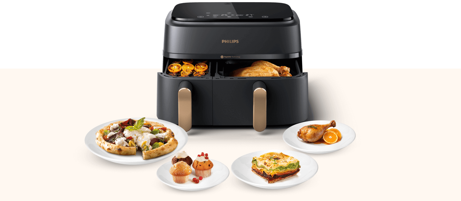 Na obrazie widoczna jest frytkownica beztłuszczowa z dwoma otwartymi koszami, w których znajdują się różne potrawy. Przed urządzeniem na talerzach umieszczone są dania, takie jak pizza, babeczki, zapiekanka i pieczone udko z kurczaka.