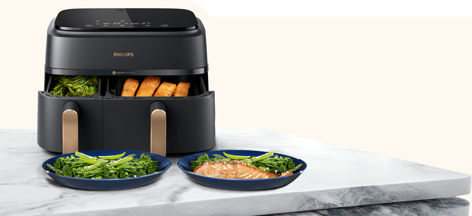 Na marmurowym blacie stoi czarna frytkownica beztłuszczowa z dwoma otwartymi koszami, w których znajdują się warzywa i pieczywo. Przed urządzeniem leżą dwa talerze z jedzeniem, a na frytkownicy widoczne jest logo \'PHILIPS\'.