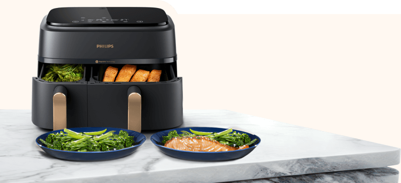 Czarna frytkownica beztłuszczowa z dwoma wysuniętymi koszami, w których znajdują się warzywa i pieczywo. Na blacie przed frytkownicą stoją dwa talerze z potrawami, a na urządzeniu widoczne jest logo \'PHILIPS\'.