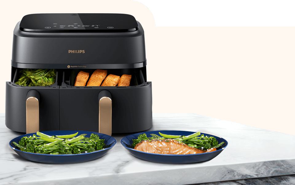 Czarna frytkownica beztłuszczowa z dwoma wysuniętymi koszami, w których znajdują się warzywa i kawałki pieczywa. Przed urządzeniem na marmurowym blacie stoją dwa talerze z zielonymi warzywami i kawałkiem łososia.