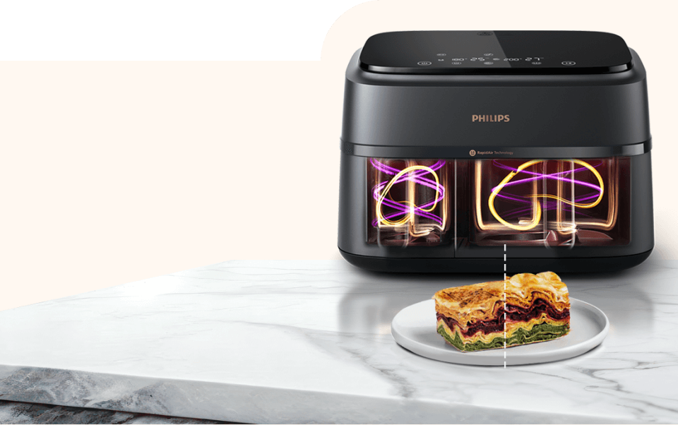 Na obrazie widoczna jest czarna frytkownica beztłuszczowa z podwójnym koszem marki PHILIPS, umieszczona na marmurowym blacie. Przed urządzeniem znajduje się talerz z kawałkiem zapiekanki.