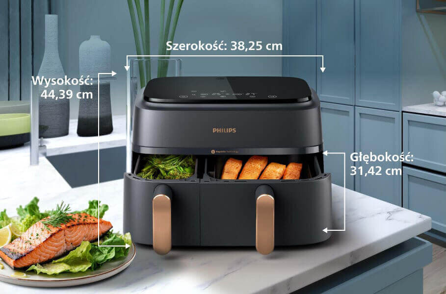 Na blacie kuchennym stoi czarna frytkownica beztłuszczowa z podwójnym koszem, z widocznym logo PHILIPS. Obok frytkownicy znajduje się talerz z łososiem i warzywami, a na urządzeniu widoczne są wymiary: szerokość 38,25 cm, wysokość 44,39 cm, głębokość 31,42 cm.