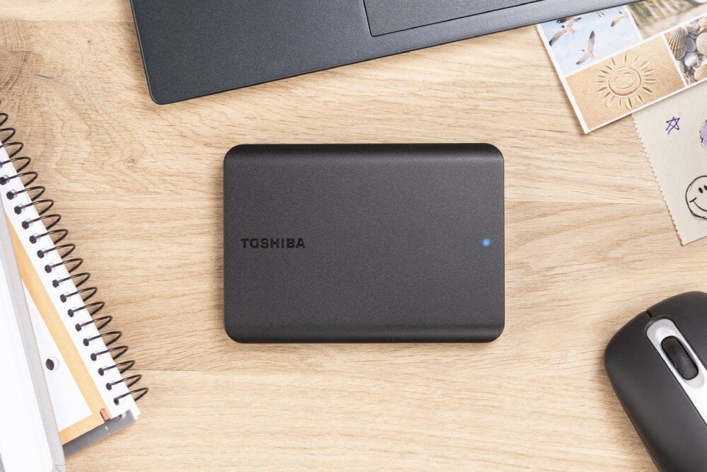 Dysk TOSHIBA Canvio Basics Exclusive Prostokątny dysk zewnętrzny leży płasko na drewnianym blacie. Pośrodku widoczne jest logo marki 'TOSHIBA', a po prawej stronie świeci się mała, niebieska dioda, opis