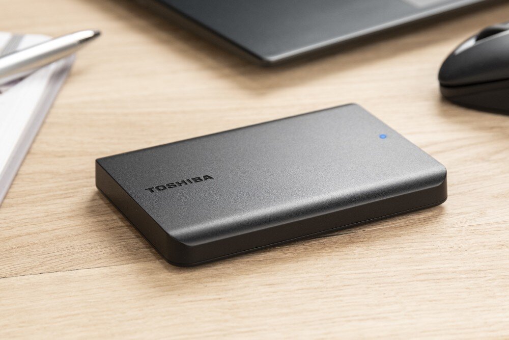 Dysk TOSHIBA Canvio Basics Exclusive Przenośny dysk twardy o matowej powierzchni spoczywa pod lekkim kątem na drewnianym biurku. Zbliżenie pokazuje logo marki i świecącą na niebiesko diodę kontrolną, pojemność, typ, obudowa