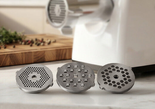 Na blacie leżą trzy wymienne sitka do maszynki do mielenia mięsa MMM-05. W tle widać urządzenie.