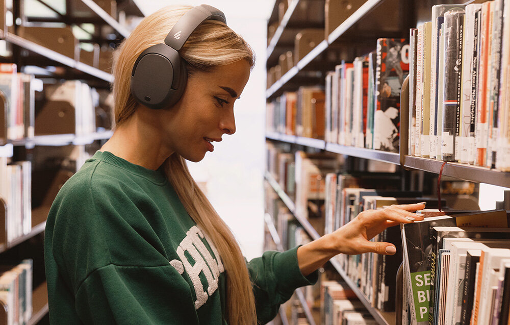 Osoba w zielonej bluzie przegląda książki na półce w bibliotece, mając na sobie szare słuchawki nauszne. W tle widać rzędy książek na regałach.