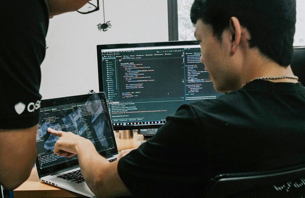 Komputer ACER Atlos BrainSphere Dwóch mężczyzn pochylonych nad laptopem i monitorem, analizujących linie kodu programistycznego w ciemnym pomieszczeniu biurowym, oprogramowanie, rozszerzanie projektów