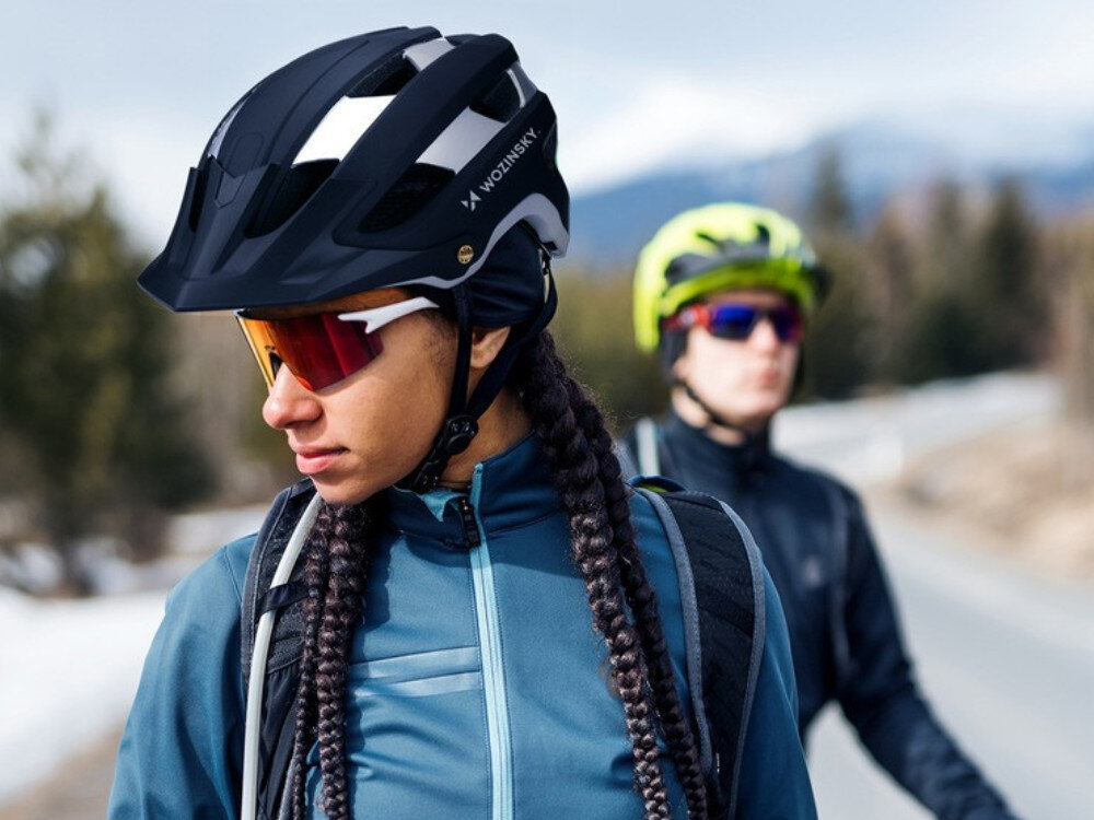 Osoba nosi czarno-srebrny kask rowerowy z napisem \'WOZINSKY\'. W tle widoczna jest druga osoba w kasku oraz rozmyty krajobraz z drzewami i górami.