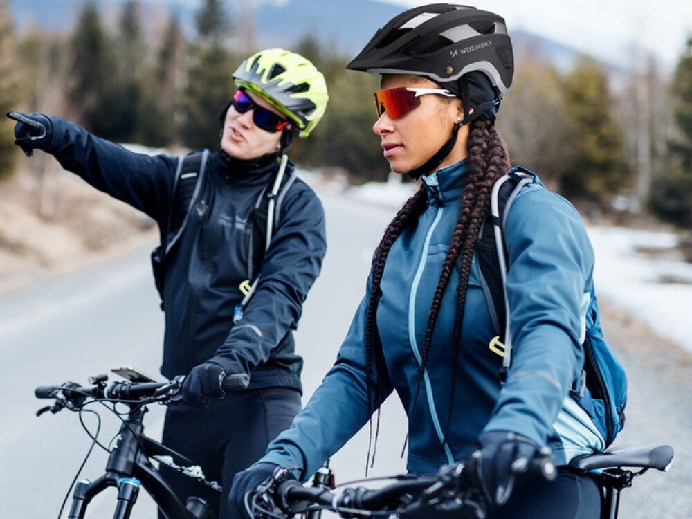 Dwie osoby na rowerach w kaskach, jedna z nich nosi czarno-srebrny kask z napisem \'WOZINSKY\'. Osoby są ubrane w sportowe kurtki i okulary przeciwsłoneczne, a w tle widać drogę i drzewa.
