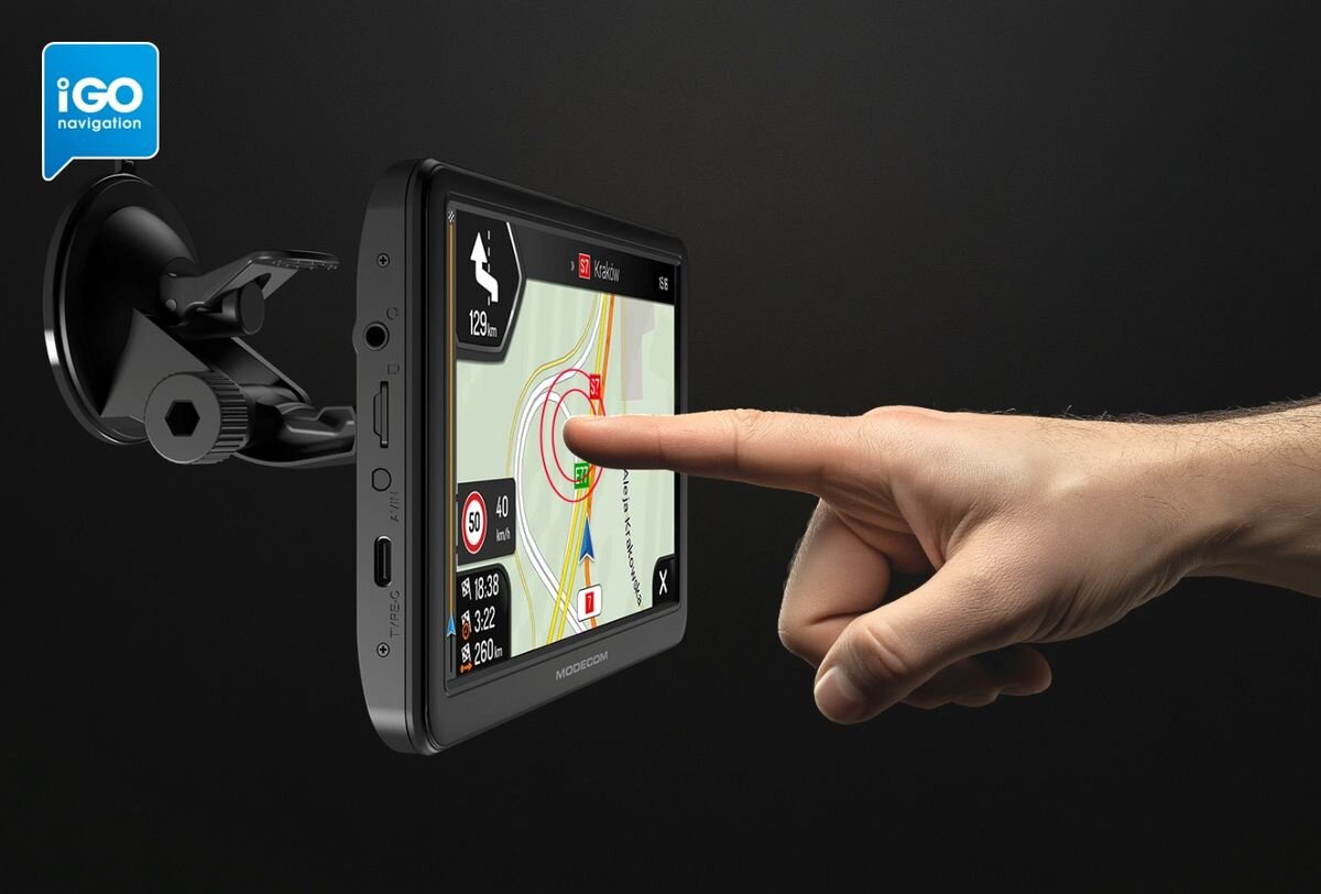 Na obrazie widoczna jest nawigacja samochodowa zamontowana na uchwycie, z wyświetlonymi mapami i danymi drogowymi. Logo \'iGO navigation\' znajduje się w lewym górnym rogu, a na ekranie widoczny jest napis \'MODECOM\'.
