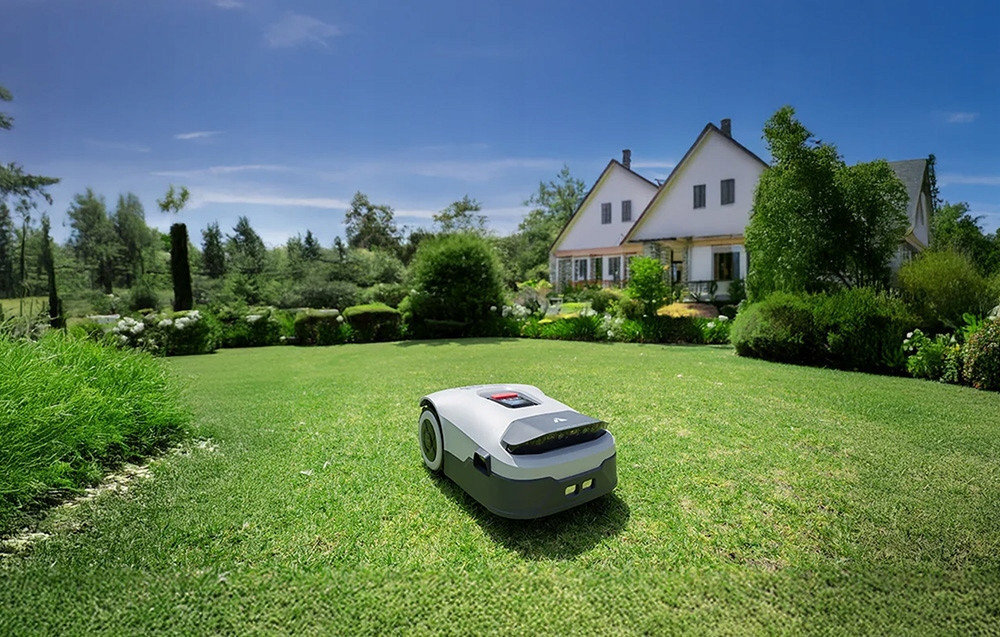 Na trawniku znajduje się robot koszący, a w tle widoczne są dwa domy otoczone zielenią. Niebo jest bezchmurne, a otoczenie jest dobrze oświetlone.
