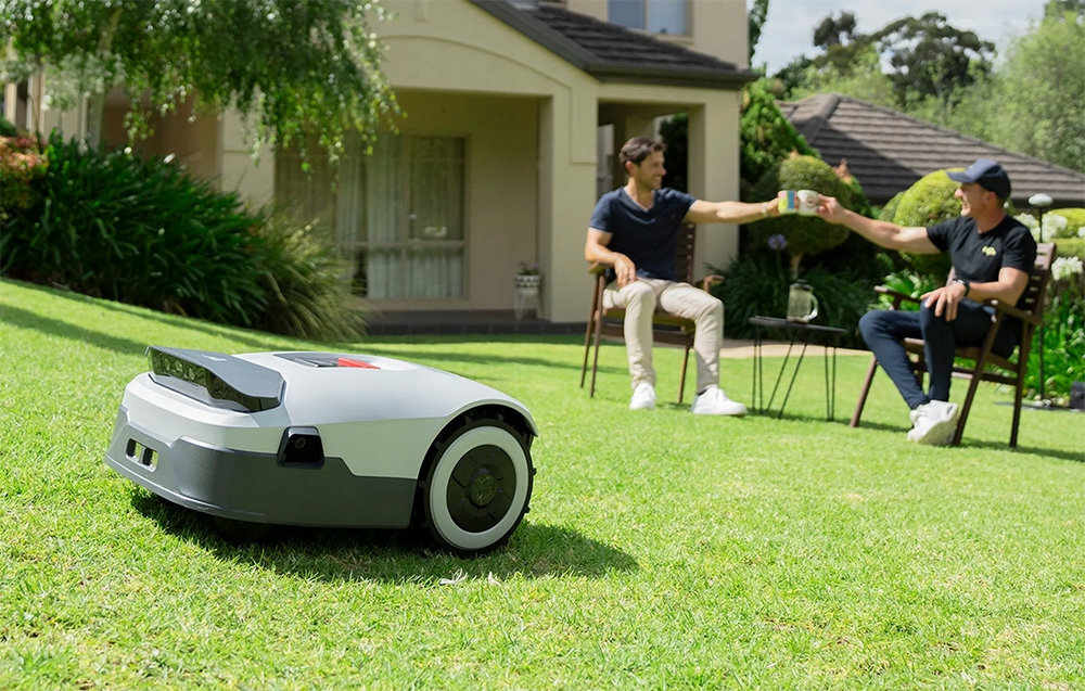 Na trawniku przed domem znajduje się robot koszący w kolorze szaro-białym. W tle dwie osoby siedzą na krzesłach i podają sobie kubek.