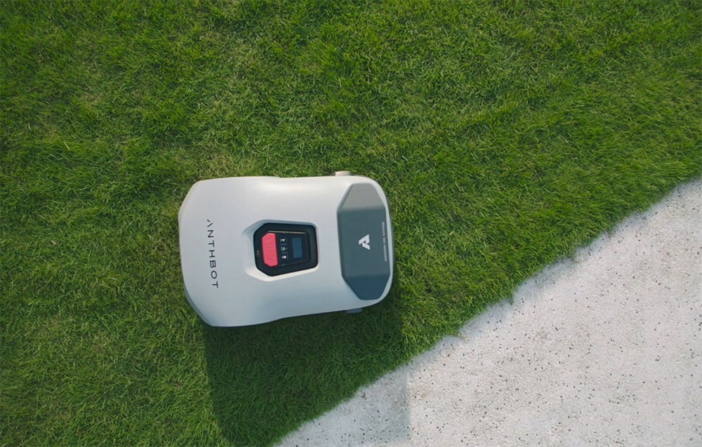 Robot koszący ANTHBOT Genie 1000 znajduje się na trawie obok betonowej powierzchni. Na obudowie widoczne jest logo w kształcie litery \'A\'.