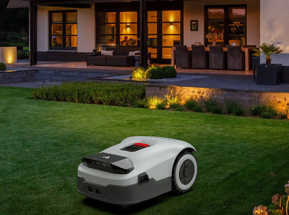Na trawniku przed domem znajduje się robot koszący o nowoczesnym designie. W tle widać oświetlony taras z meblami ogrodowymi.