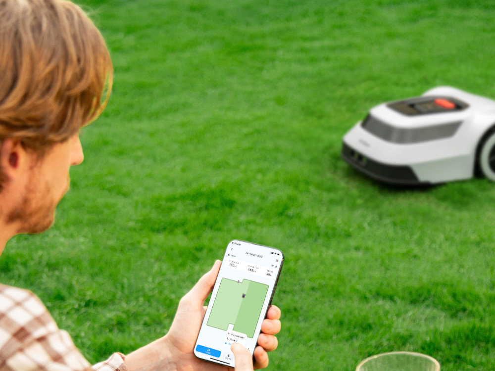 Robot koszący ANTHBOT Genie 3000 Bez przewodu ograniczającego ogród trawnik robot stoi na trawie na pierwszym planie osoba z telefonem w dłoni łączność Wi-Fi i aplikacja Anthbot App dokładny harmonogram koszenia na konkretne dni i godziny zmienisz tryb pracy sprawdzisz aktualny status urządzenia w czasie rzeczywistym kontrolować postęp koszenia i reagować