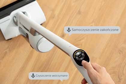Osoba trzyma odkurzacz na drewnianej podłodze, z widocznymi komunikatami: \'Samoczyszczenie zakończone\' i \'Czyszczenie wstrzymane\'.