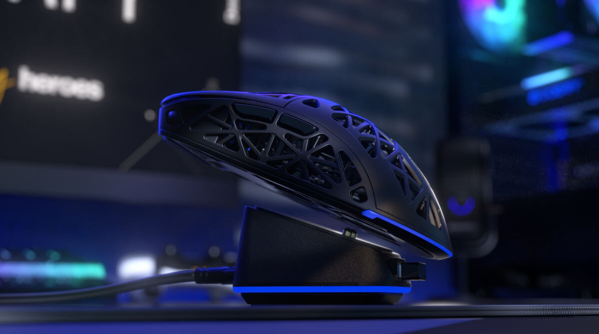 Na obrazie widoczna jest czarna mysz komputerowa z ażurową obudową, umieszczona na stacji ładującej z niebieskim podświetleniem. W tle znajduje się rozmyte wnętrze z elementami komputerowymi.