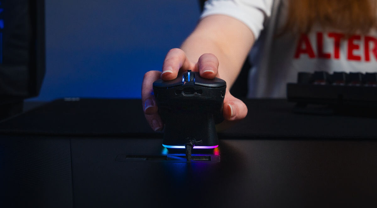 Osoba trzyma mysz komputerową z podświetleniem RGB na ciemnym biurku. W tle widoczna jest klawiatura i fragment koszulki z napisem \'ALTER\'.