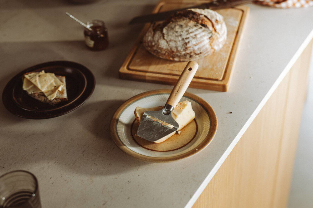 Na blacie kuchennym znajduje się krajalnica do sera na talerzu z kawałkiem sera. Obok leży chleb na desce do krojenia oraz słoik z dżemem i talerz z kromkami chleba.