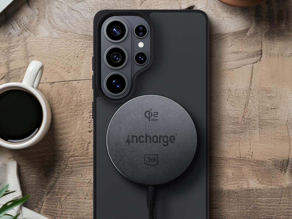 Na drewnianym stole leży smartfon z podłączoną czarną ładowarką indukcyjną z widocznymi napisami \'qi2 25W\', \'incharge\' i logo \'3mk\'. Obok znajduje się filiżanka z czarną kawą.
