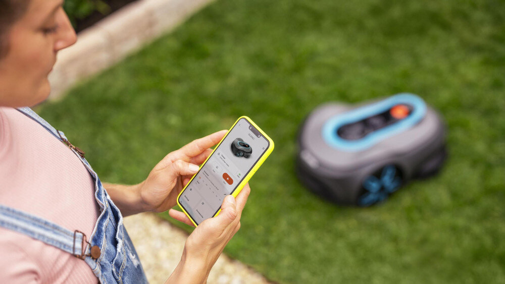 Robot koszący GARDENA Smart Sileno Free 600 19921-32 sterowanie Wi-Fi robot stoi na trawie osoba kuca obok z telefonem w dłoni tworzenie harmonogramu koszenia aktualizacje realizowane bezprzewodowo dzięki funkcji FOTA moduł Bluetooth łączność z aplikacją GARDENA Bluetooth® App lub GARDENA Smart App na systemy iOS 17/Android 9 lub nowsze