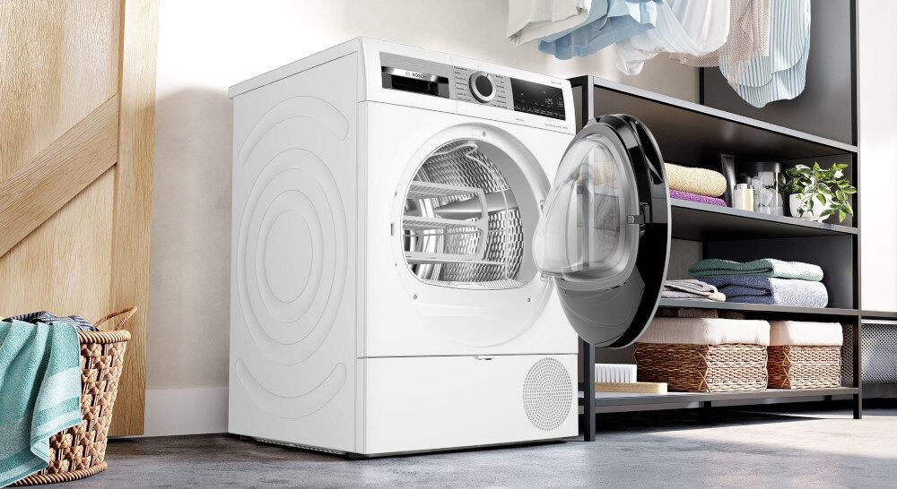 SUSZARKA BOSCH WQG241MSPL Na zdjęciu widać suszarkę bębnową z otwartymi drzwiami. Urządzenie znajduje się w pralni lub garderobie. Po lewej stronie umieszczony jest kosz z ręcznikiem, a po prawej regał z poukładanymi tekstyliami, koszami oraz rośliną. Nad regałem wiszą ubrania. Wnętrze suszarki jest puste, przygotowane do załadunku. Całość wygląda czysto i nowocześnie cicha praca suszarki bosch redukcja hałasu antywibracje antivibration design stabilizacja bębna komfortowe suszenie minimalne drgania izolacja ścian amortyzator