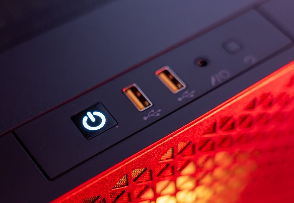 Zbliżenie na panel przedni komputera z podświetlanym przyciskiem zasilania oraz dwoma portami USB. Obudowa ma czerwone podświetlenie i wzór z trójkątnymi otworami.