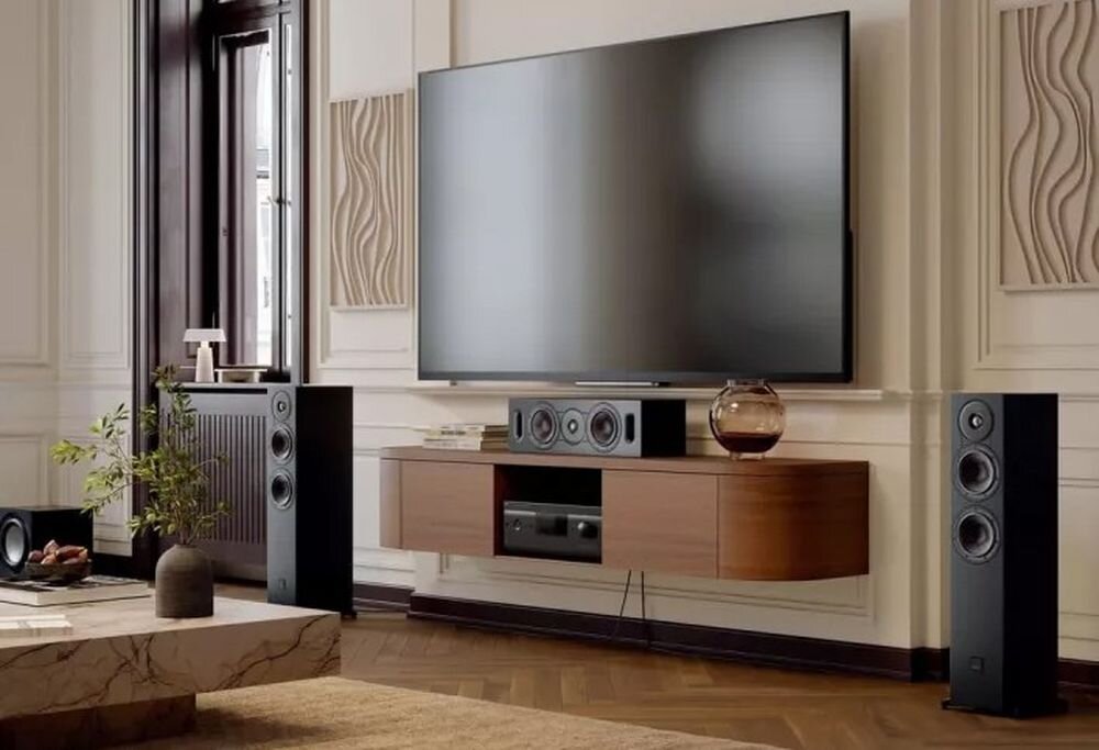 W eleganckim salonie znajduje się duży telewizor zamontowany na ścianie, poniżej którego stoi drewniana szafka z głośnikiem centralnym i sprzętem audio. Po obu stronach szafki umieszczone są wysokie głośniki, a na stoliku obok stoi roślina w doniczce.