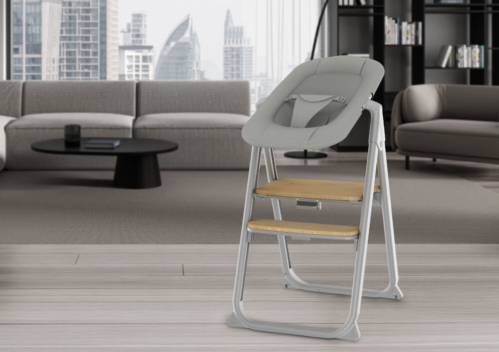 Szare krzesełko do karmienia z drewnianymi elementami stoi w nowoczesnym salonie z sofą i stolikiem kawowym. W tle widoczne są wysokie budynki za oknem.