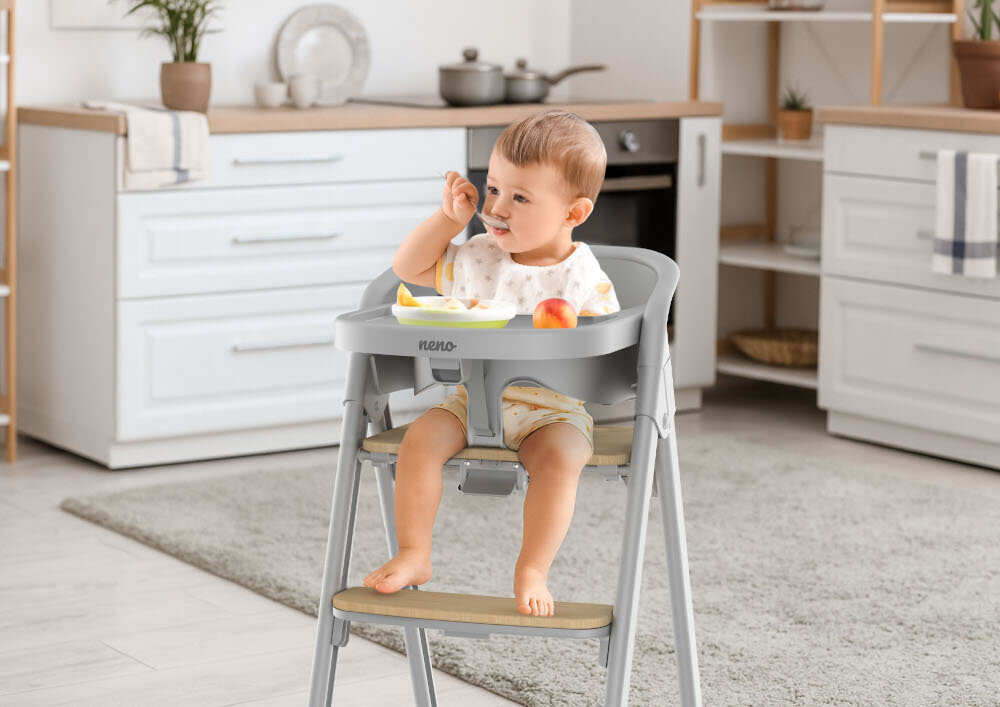 Dziecko siedzi w szarym krzesełku do karmienia z napisem \'neno\' na tacy. W tle widoczna jest jasna kuchnia z białymi szafkami i drewnianymi blatami.