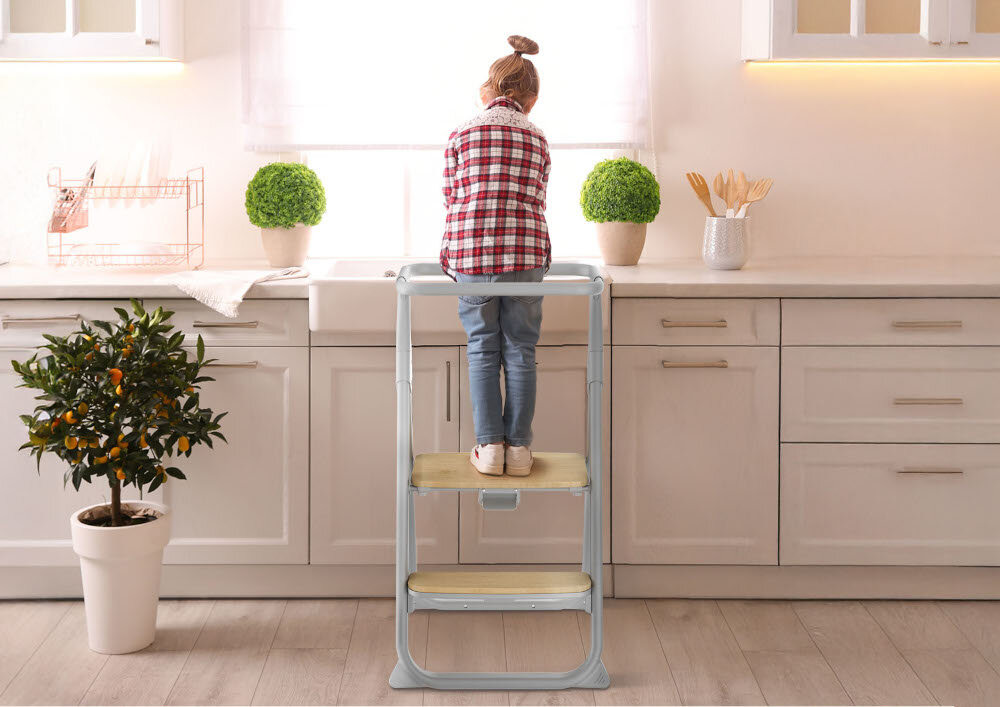 Dziecko stoi na szarym krzesełku do karmienia przy kuchennym blacie. W tle widać białe szafki, rośliny doniczkowe i akcesoria kuchenne.