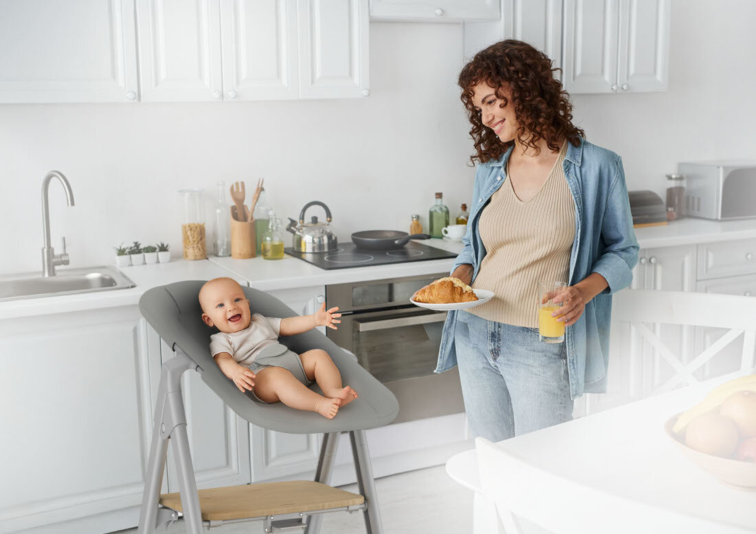 W jasnej kuchni stoi szare krzesełko do karmienia z uśmiechniętym dzieckiem. Obok osoba trzyma talerz z rogalikiem i szklankę soku pomarańczowego.