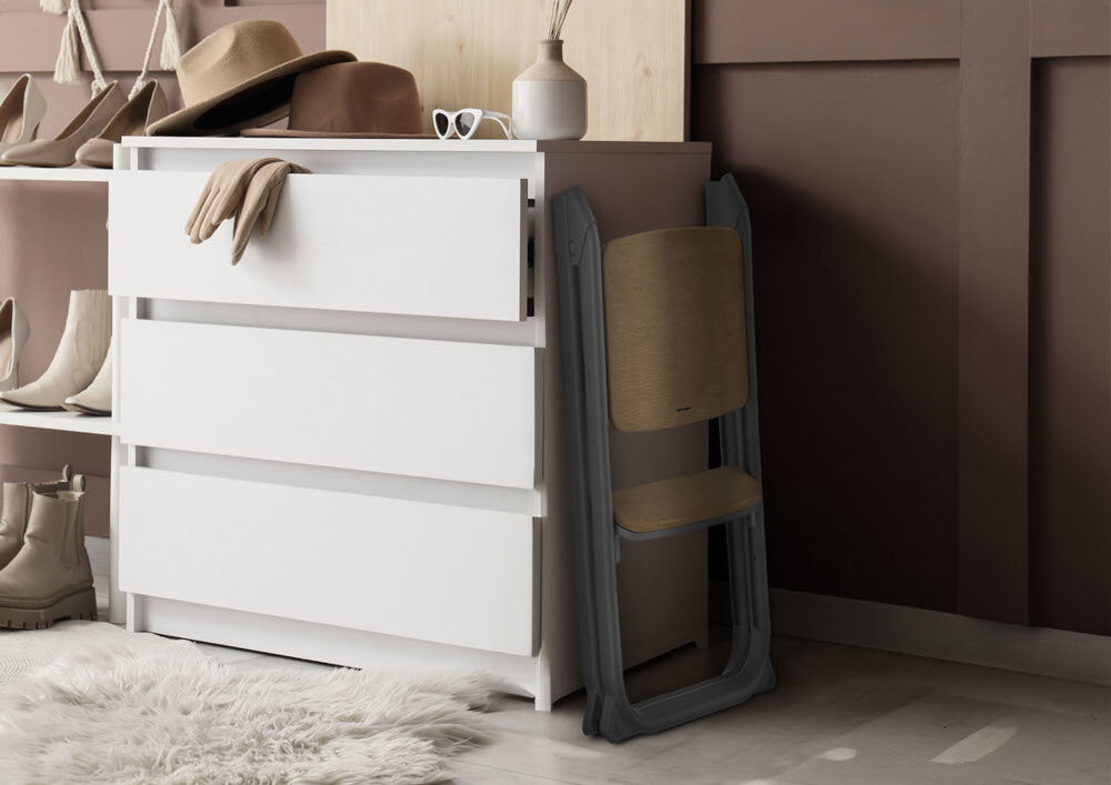 Wnętrze z białą komodą, na której znajdują się kapelusz, okulary i wazon. Obok komody stoi złożone krzesełko w szarym kolorze.