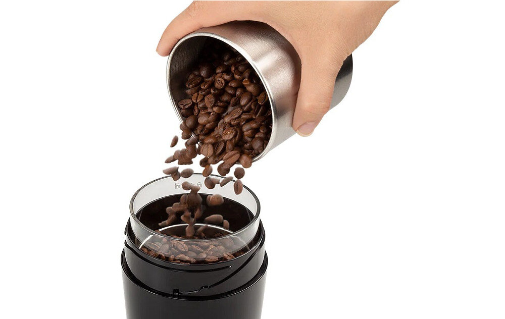 Osoba wsypuje ziarna kawy z metalowego pojemnika do młynka do kawy. Młynek jest czarny i ma przezroczysty pojemnik na ziarna.