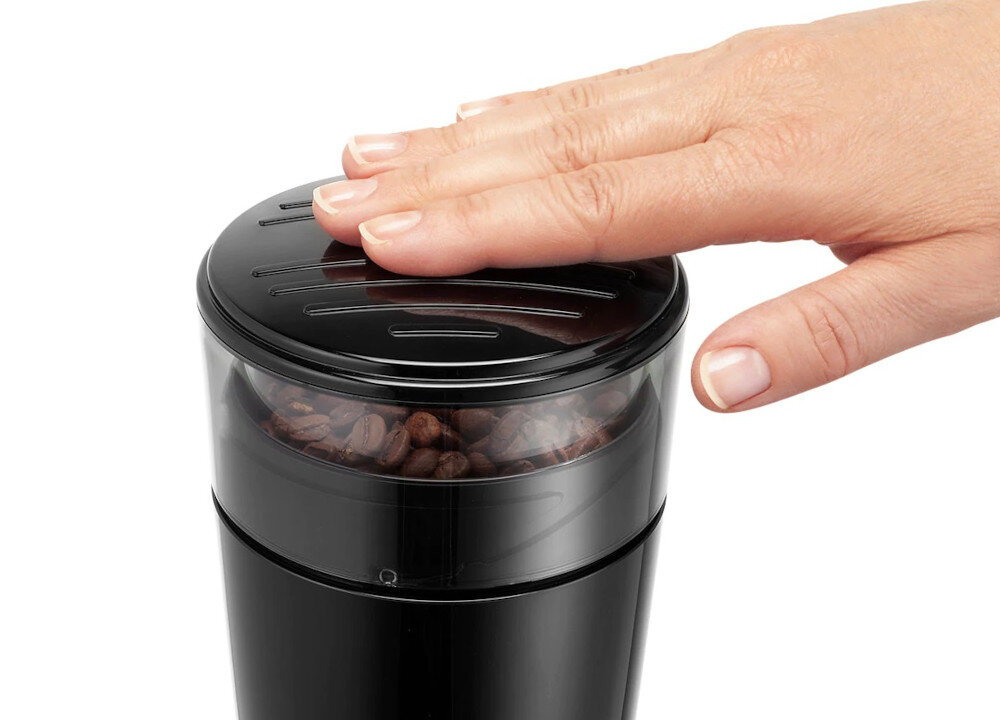 Czarna obudowa młynka do kawy z przezroczystą pokrywą, pod którą widać ziarna kawy. Na pokrywie spoczywa dłoń.