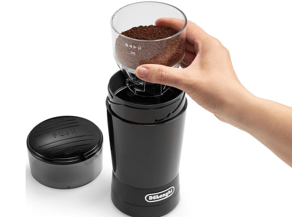 Czarny młynek do kawy z logo \'DeLonghi\' na obudowie, obok znajduje się pokrywka z napisem \'PUSH\'. Ręka trzyma przezroczysty pojemnik z mieloną kawą nad młynkiem.