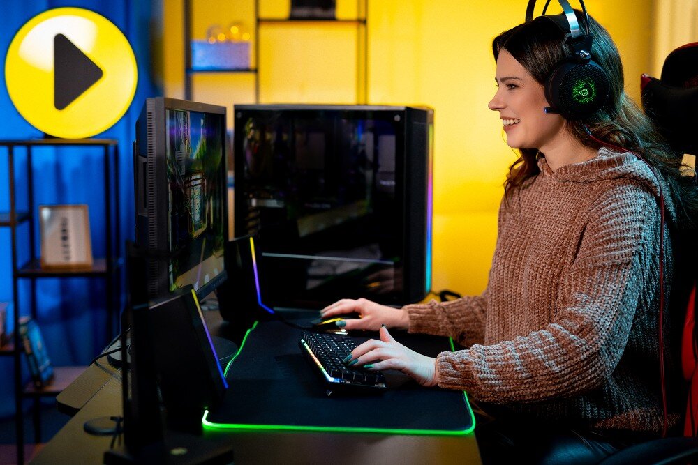 Komputer MAD DOG GeForce RTX5090 DLSS 4 Reflex Edition 2 BQ900FX-A02WR64WH Uśmiechnięta kobieta w słuchawkach siedzi przy biurku z komputerem, podświetlaną klawiaturą i myszką. Obok monitora stoi czarna obudowa z przeszklonym bokiem, a w tle widoczne jest dekoracyjne oświetlenie oraz żółta ikona 'play'